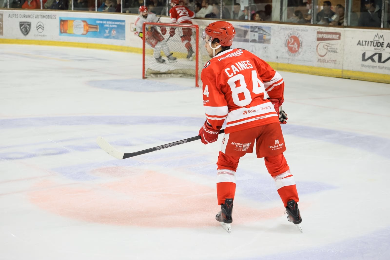 Harrison Caines, n° 84 des Diables Rouges, de dos en maillot rouge, crosse en main sur la glace de René-Froger, devant la cage et les panneaux des partenaires