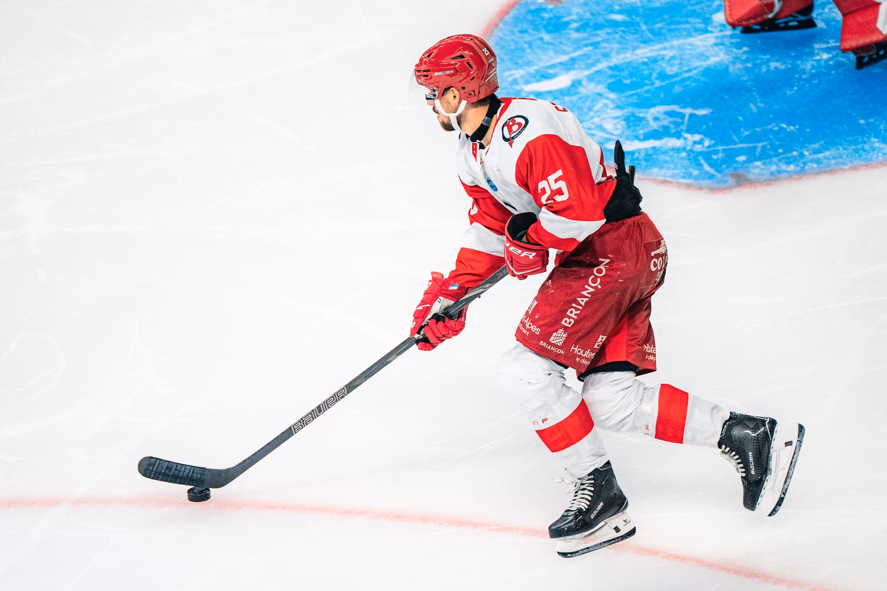 Un joueur des Diables Rouges, casque rouge et maillot blanc, conduit le palet sur la glace, la crosse tendue devant lui ; derrière, le gardien briançonnais devant sa zone