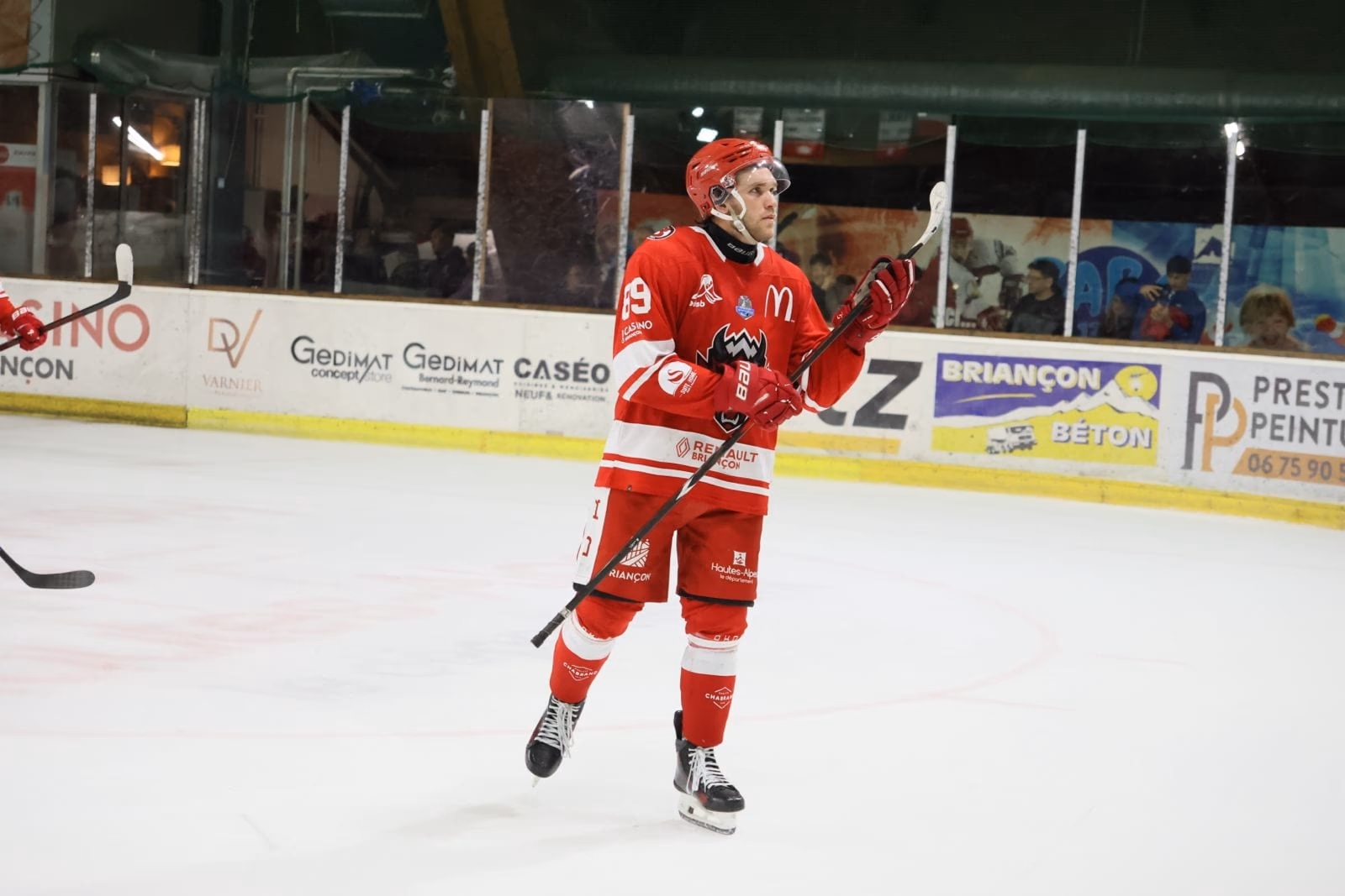 Un joueur des Diables Rouges en maillot rouge, crosse à la main, sur la glace de René-Froger devant les panneaux des partenaires