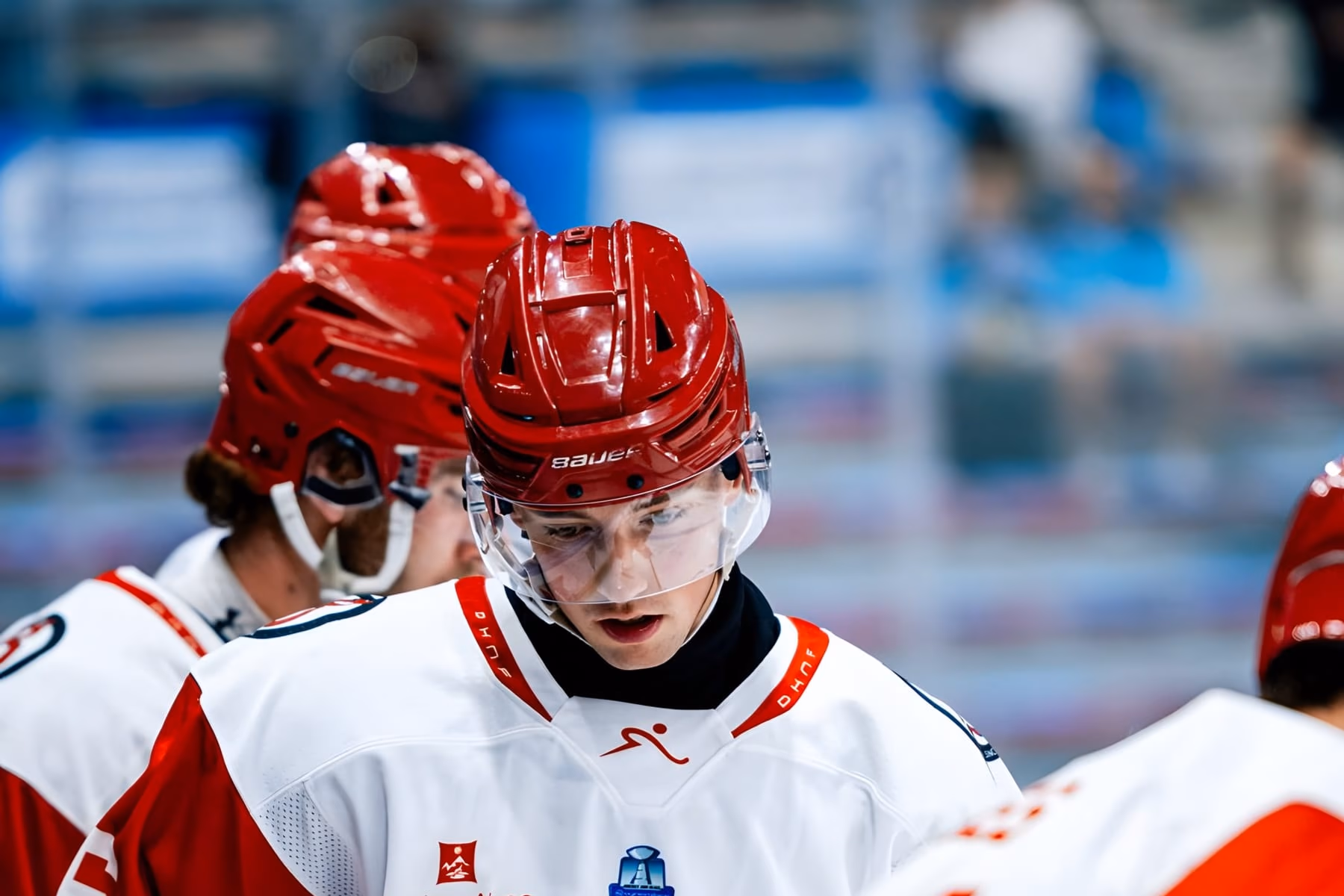 Un joueur des Diables Rouges tête baissée, casque rouge et maillot blanc, au milieu de ses coéquipiers ; au premier plan, les gourdes rouges du banc